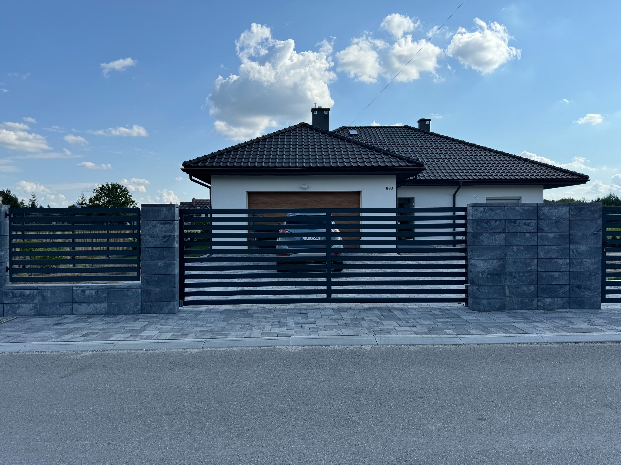 Nowoczesna, pozioma brama wjazdowa z szarymi, betonowymi słupkami na tle białego domu z ciemnym dachem i błękitnego nieba z chmurami.