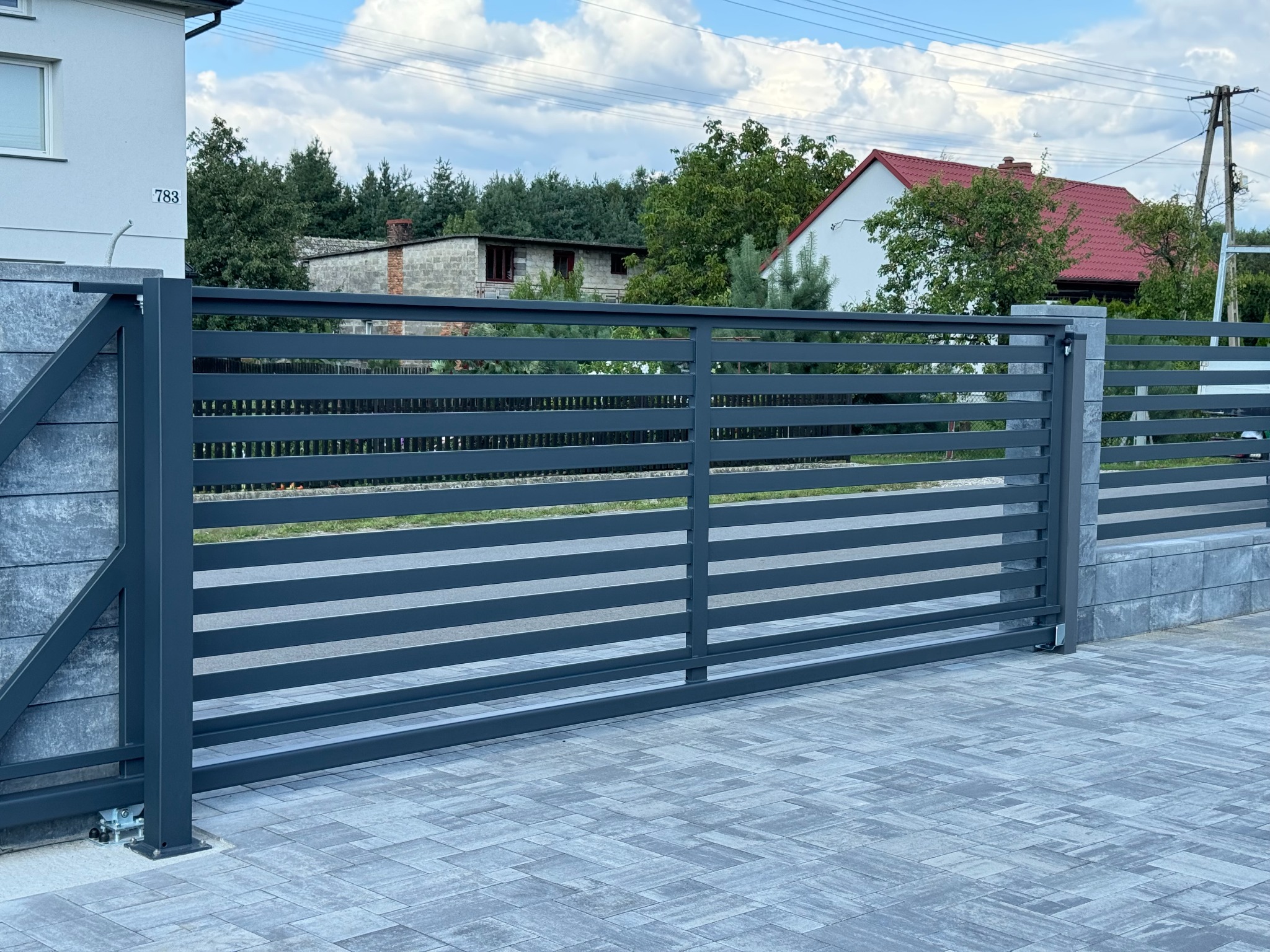 Nowoczesna, przesuwna brama wjazdowa w kolorze antracytowym z poziomymi panelami, zamontowana na posesji z szarej kostki brukowej i betonowych słupków.