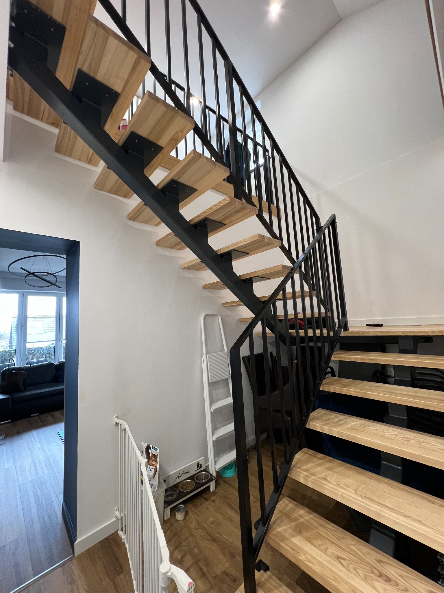 Nowoczesne schody z drewnianymi stopniami i czarną metalową konstrukcją, widok z dołu, minimalistyczne wnętrze z białymi ścianami i drewnianą podłogą, barierka zabezpieczająca dla dzieci na dole...