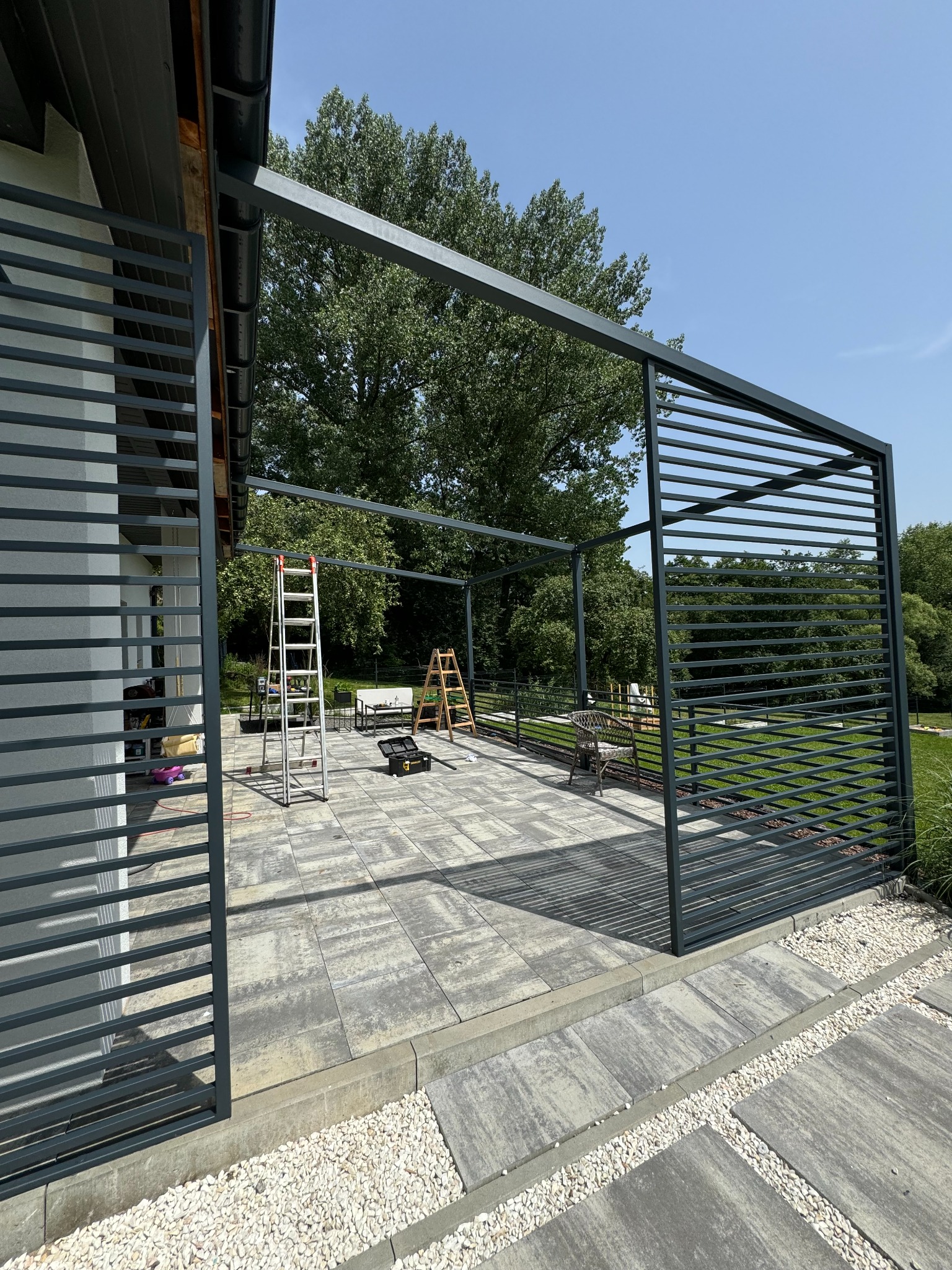 Stalowa konstrukcja pergoli w trakcie montażu na tarasie z szarych płyt, widoczne drabiny i narzędzia, w tle trawnik i drzewa.