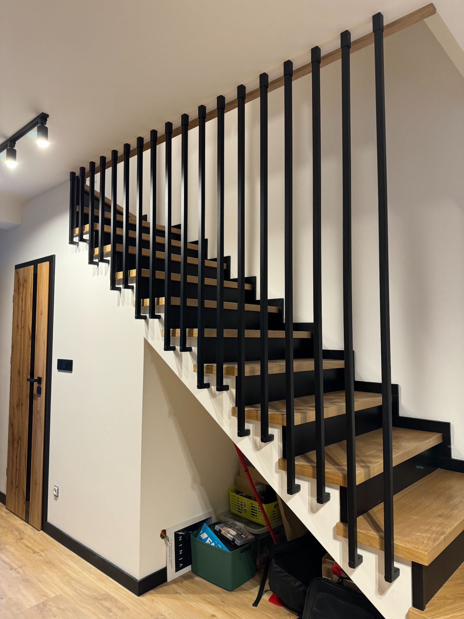 Nowoczesne schody drewniane z czarną, minimalistyczną balustradą, widok z dołu, oświetlenie punktowe, drzwi z drewnianą okleiną w tle.