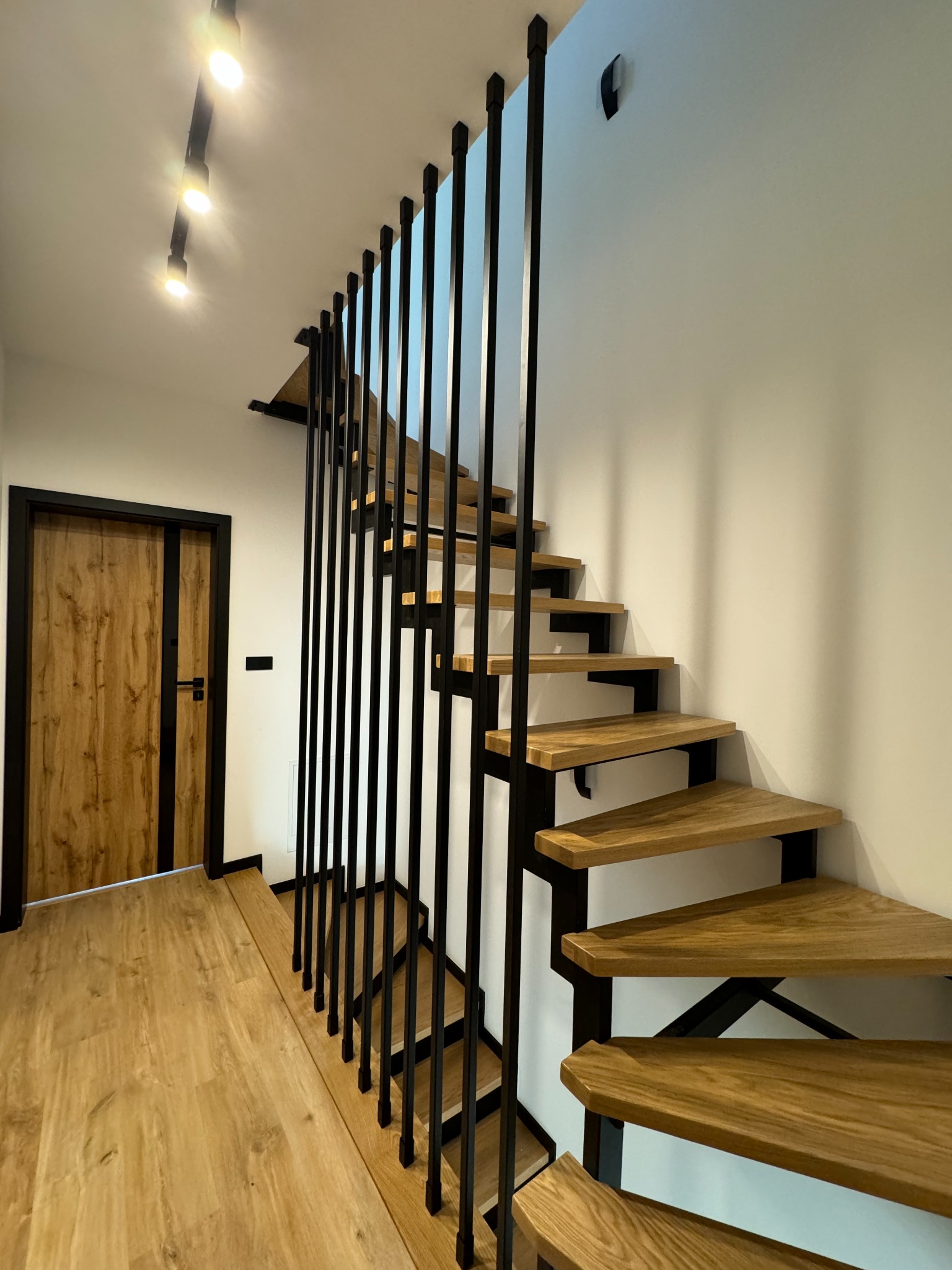 Nowoczesne schody drewniane z czarną, pionową balustradą, oświetlenie punktowe sufitowe, widok z dołu na górę, drzwi w tle.