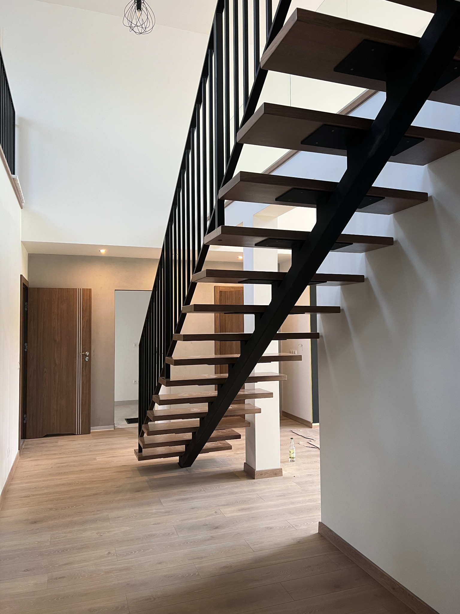 Nowoczesne schody z drewnianymi stopniami i czarną metalową konstrukcją w jasnym wnętrzu domu, widok z dołu na otwartą przestrzeń z drewnianą podłogą i drzwiami.