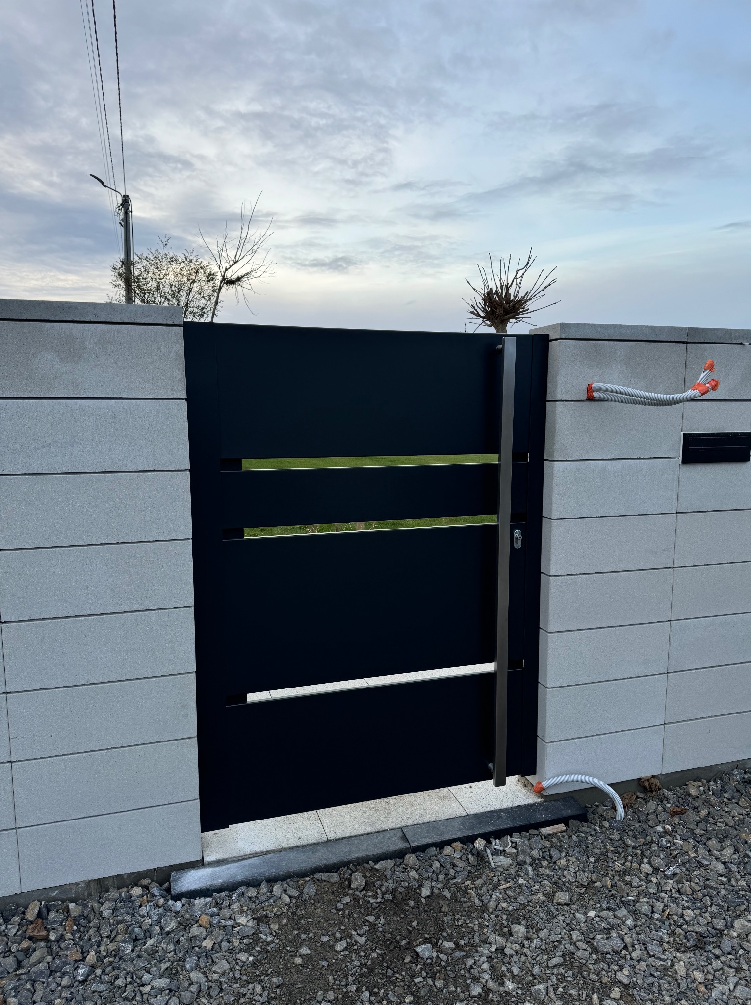 Nowoczesna, czarna brama wjazdowa z poziomymi szczelinami wkomponowana w szary, betonowy mur. Widoczne przyłącza elektryczne w peszlu oraz skrzynka na listy. Podłoże z szarego żwiru.