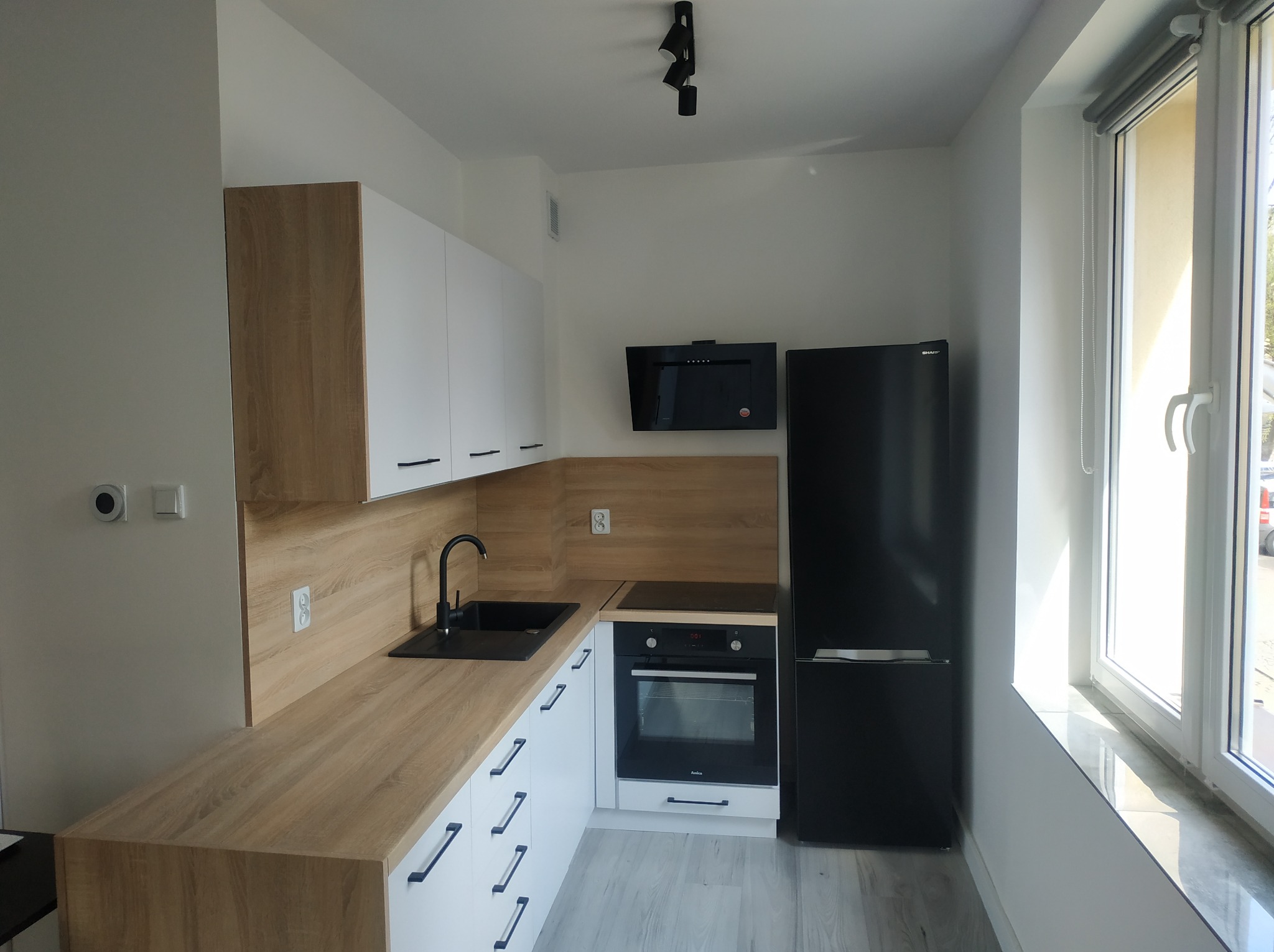 Nowoczesna, jasna kuchnia z białymi szafkami, drewnianym blatem, czarnym zlewem i armaturą, oraz czarną lodówką w zabudowie, oświetlona trzema reflektorami punktowymi.