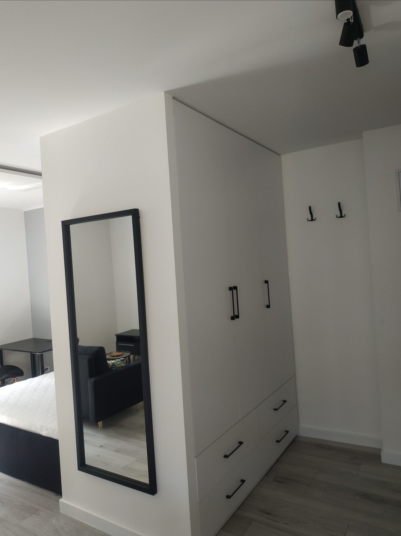 Biała szafa z czarnymi uchwytami, wbudowana w ścianę, z lustrem obok, odbijającym fragment pokoju z łóżkiem i sofą.