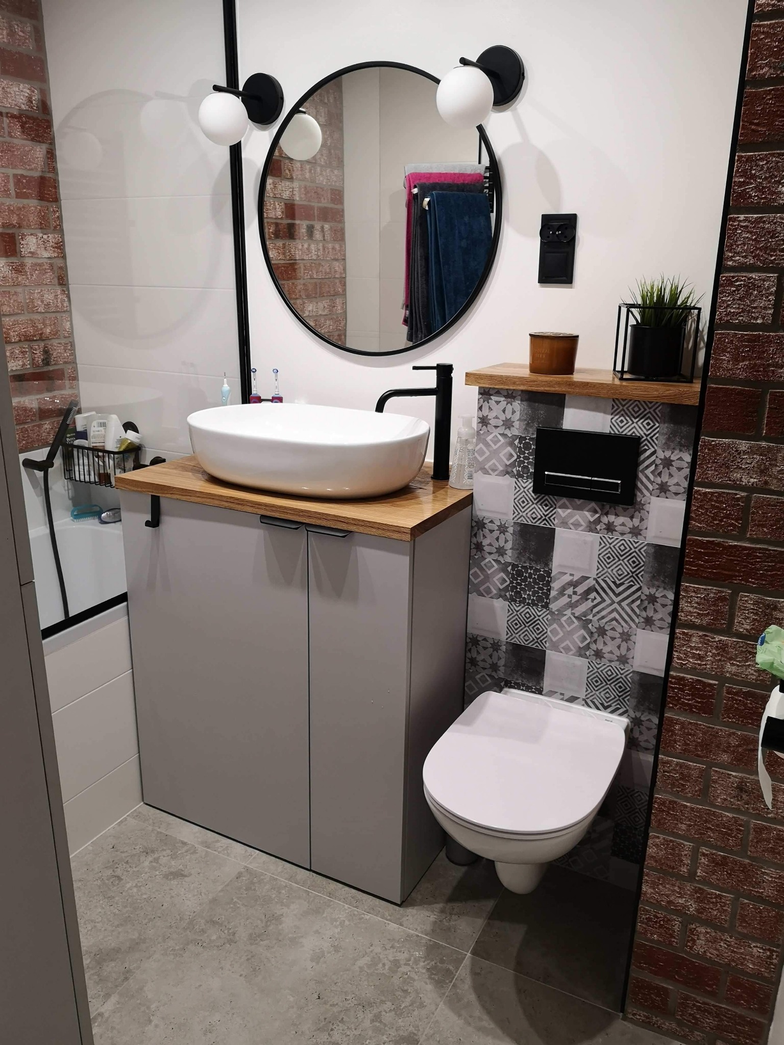 Nowoczesna łazienka z owalną umywalką nablatową na szarej szafce, okrągłe lustro z czarną ramą, ściana z cegły i mozaiki, podwieszana toaleta.