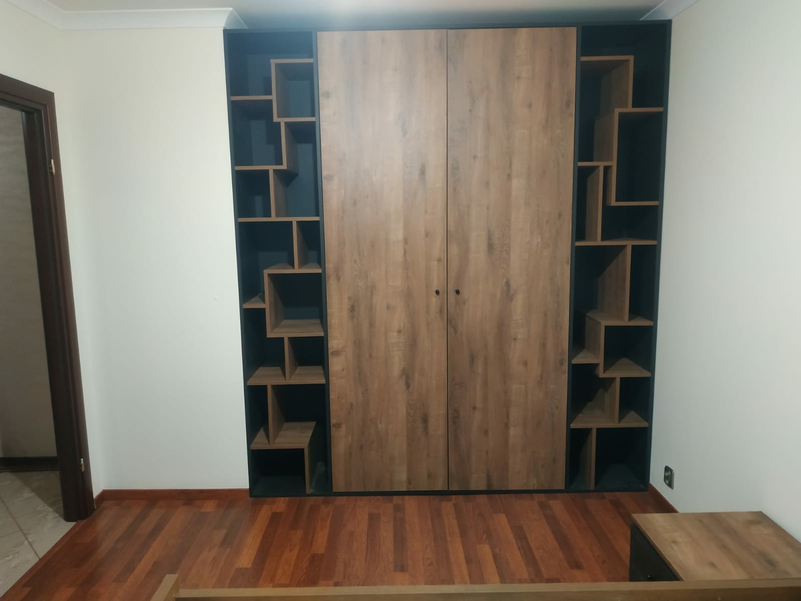 Dwudrzwiowa szafa z drewnopodobnymi frontami, flankowana asymetrycznymi regałami w kolorze grafitowym, na tle białej ściany i parkietu z brązowego drewna.