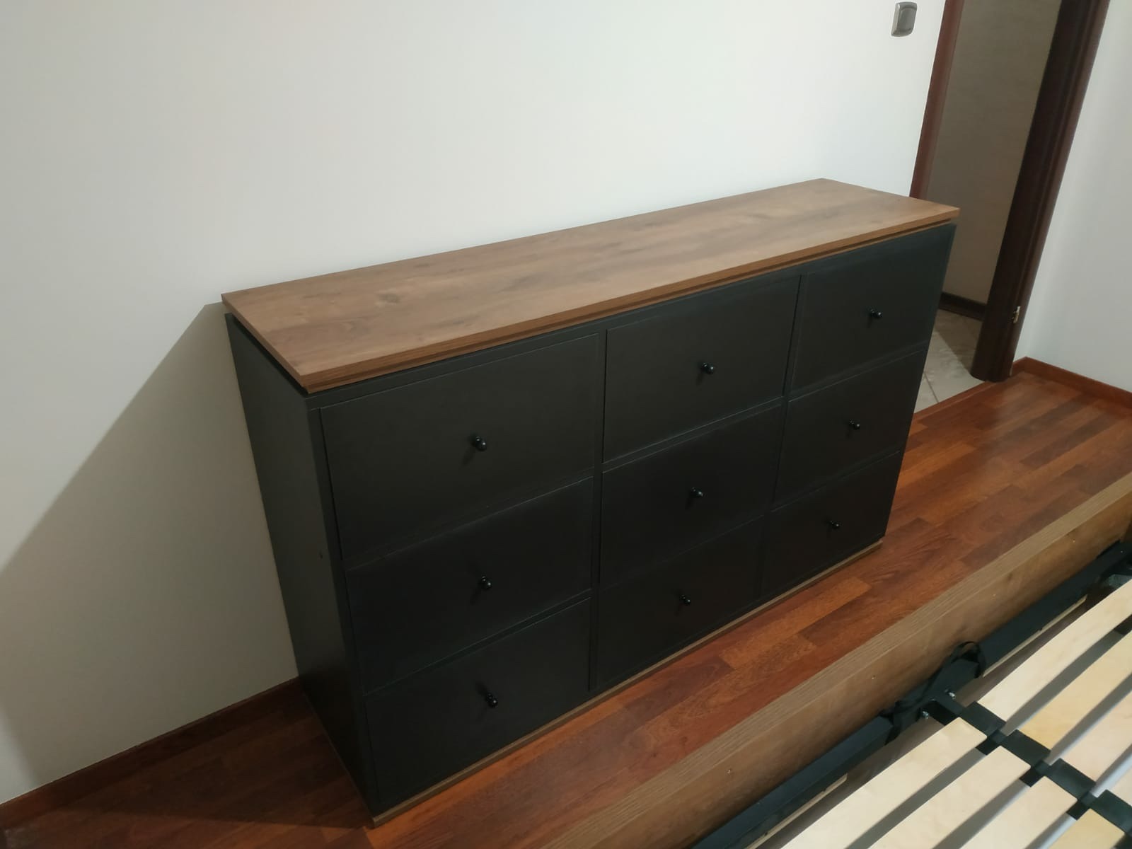 Czarna komoda z dziewięcioma szufladami i blatem w kolorze drewna, ustawiona na drewnianej podłodze przy białej ścianie, widoczny fragment ramy łóżka.