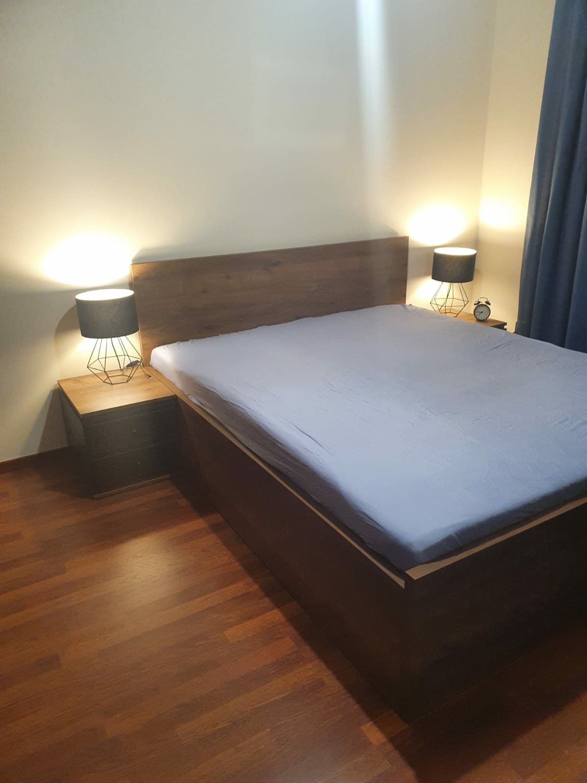 Nowoczesne łóżko z drewnianym wezgłowiem i szafkami nocnymi z geometrycznymi lampkami, oświetlone ciepłym światłem, na parkiecie.