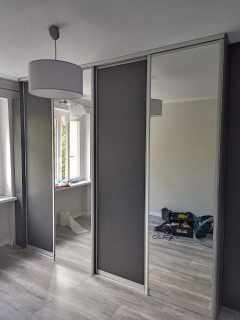 Trzydrzwiowa szafa przesuwna z szarymi panelami i lustrzanymi drzwiami, widoczne narzędzia w odbiciu lustrzanym, jasne panele podłogowe