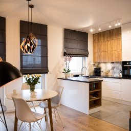 Profi Domo - Nowoczesna kuchnia z wyspą, białymi szafkami i drewnianymi akcentami, geometryczne płytki nad blatem, stół z krzesłami i lampą w stylu industrialnym, rolety rzymskie w oknach.