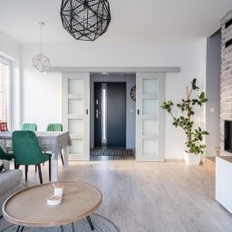 Profi Domo - Jasne, przestronne wnętrze z szarym narożnikiem, okrągłym stolikiem kawowym z rattanu, zielonymi krzesłami przy stole jadalnym i przesuwnymi drzwiami do przedpokoju, oświetlone geometrycznymi lampami.