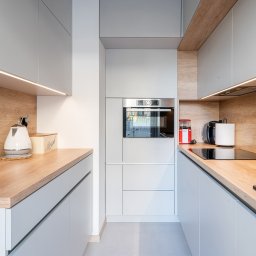 Profi Domo - Wąska kuchnia w odcieniach szarości z drewnianymi akcentami, oświetleniem LED pod szafkami i sprzętami AGD w zabudowie.