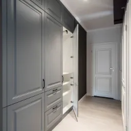 Szara szafa wnękowa z frezowanymi frontami, czarnymi uchwytami i otwartymi drzwiami ukazującymi białe półki, na tle jasnej podłogi i ciemnej ściany.