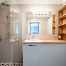 Nowoczesna łazienka z drewnianymi akcentami: kabina prysznicowa ze szklaną ścianką, armatura w kolorze chromu, umywalka nablatowa na drewnianym blacie, szafki w odcieniach szarości, płytki...