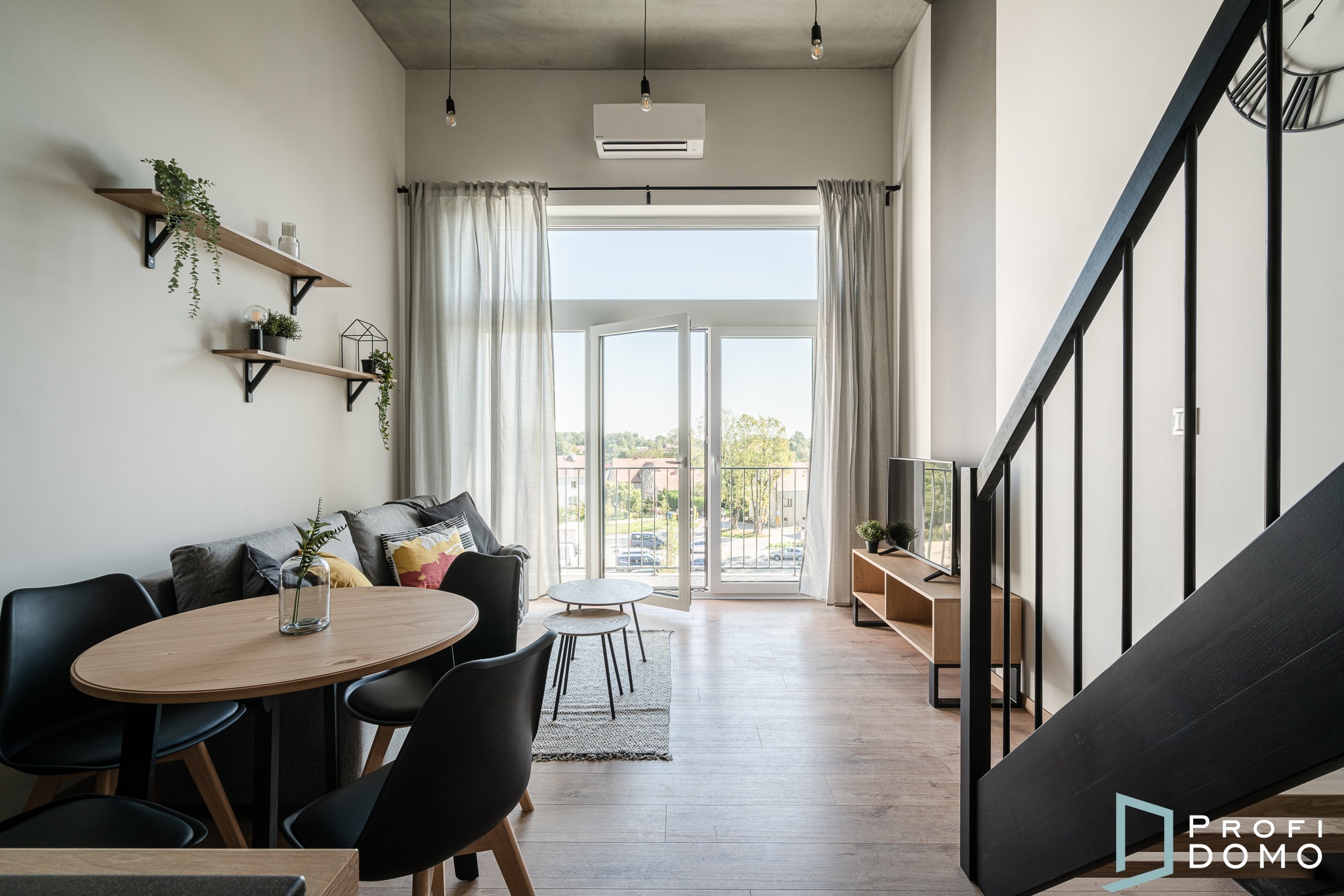 Jasne wnętrze mieszkania z drewnianą podłogą, oknem balkonowym z widokiem na zieleń, drewnianym stołem z czarnymi krzesłami, szarą sofą z poduszkami i czarnymi schodami z logo firmy.