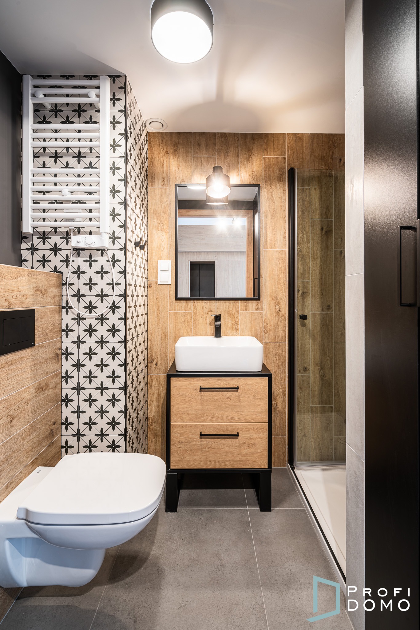 Nowoczesna łazienka z drewnopodobnymi płytkami, wiszącą toaletą, umywalką nablatową na szafce z szufladami i grzejnikiem drabinkowym na ścianie w geometryczne wzory.