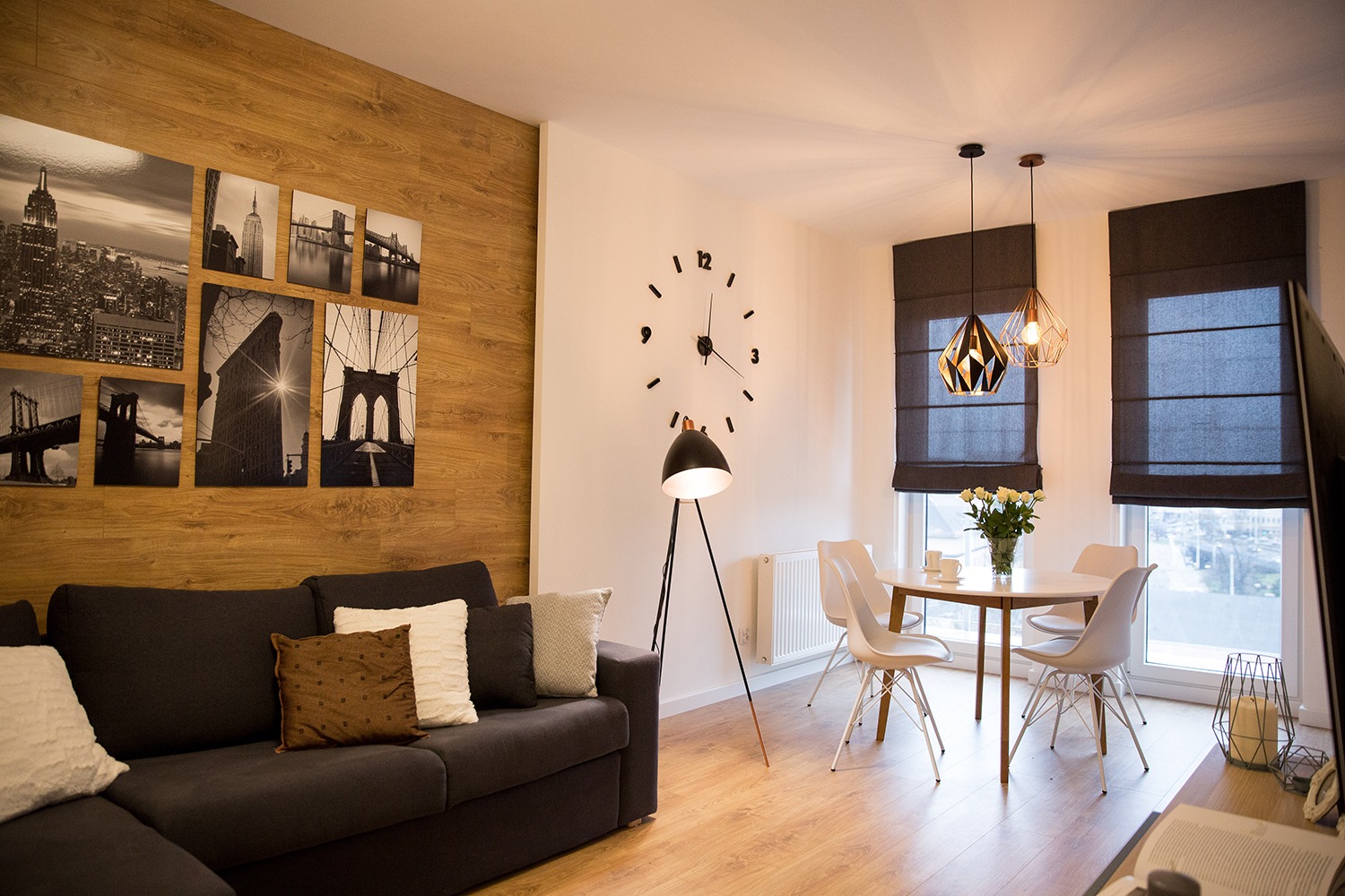 Salon z drewnianą ścianą ozdobioną czarno-białymi fotografiami Nowego Jorku, szary narożnik z poduszkami, okrągły stół z białymi krzesłami i bukietem białych róż, geometryczne lampy wiszące i duży...