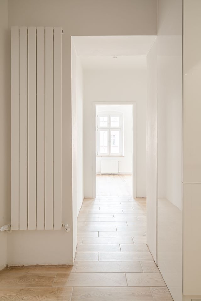 Jasny korytarz w minimalistycznym stylu z białym grzejnikiem panelowym i podłogą wyłożoną płytkami imitującymi drewno, widok na pokój z oknem.
