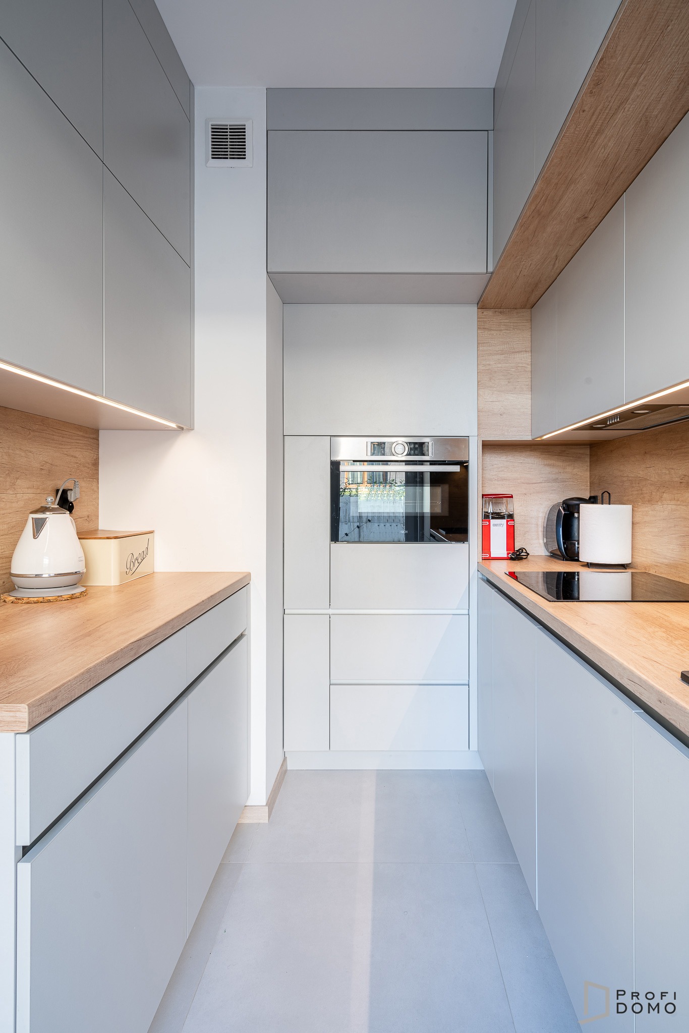 Wąska kuchnia w odcieniach szarości z drewnianymi akcentami, oświetleniem LED pod szafkami i sprzętami AGD w zabudowie.