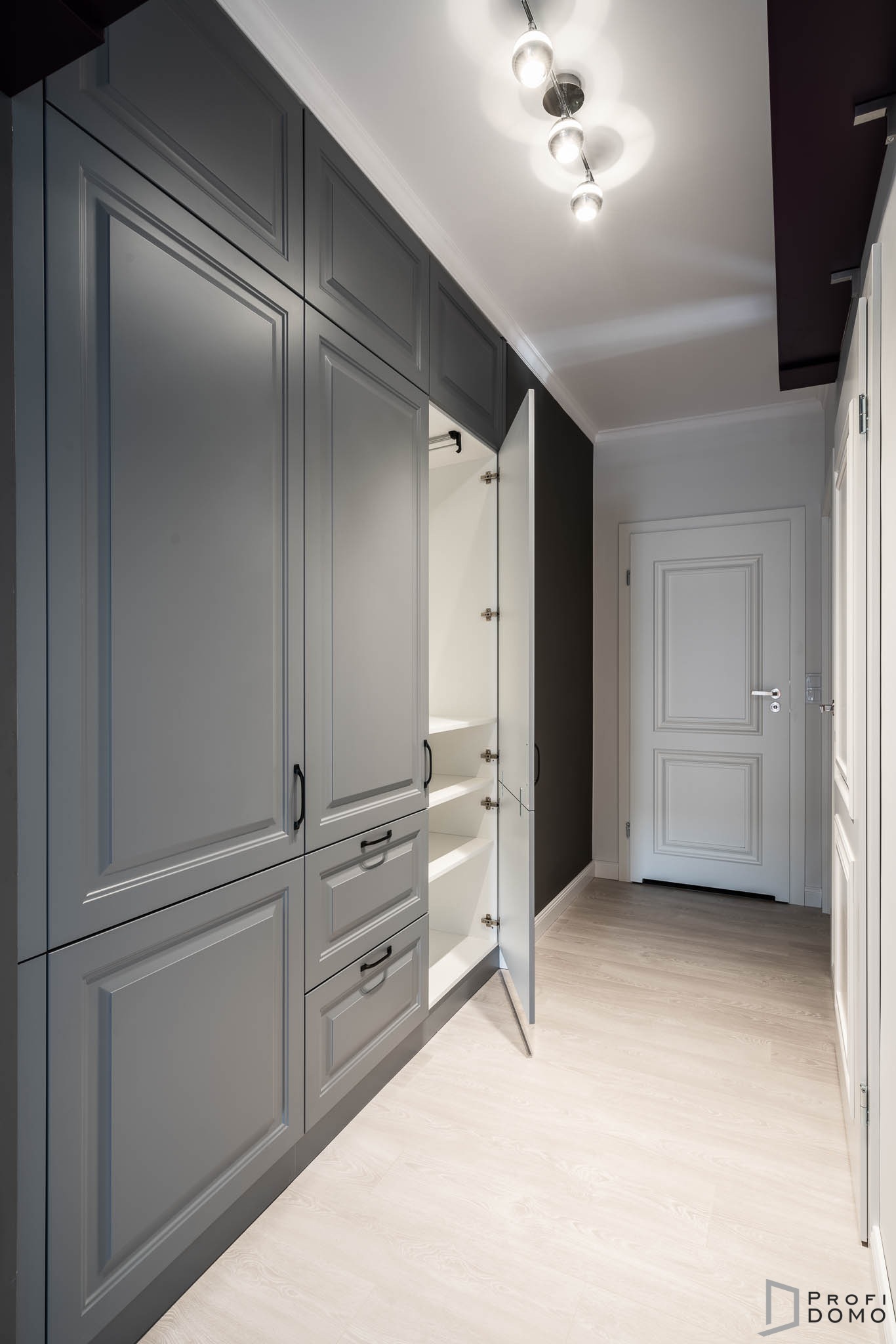 Szara szafa wnękowa z frezowanymi frontami, czarnymi uchwytami i otwartymi drzwiami ukazującymi białe półki, na tle jasnej podłogi i ciemnej ściany.