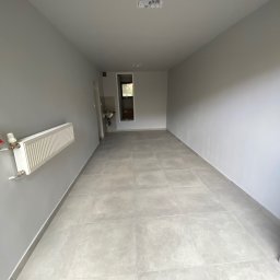 INSIDE HOME - DAMIAN RUSINEK - Pomalowane na szaro wnętrze z płytkami podłogowymi, grzejnikiem na ścianie i widocznym fragmentem łazienki w tle.