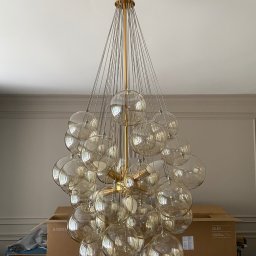 INSIDE HOME - DAMIAN RUSINEK - Złoty żyrandol z kulistymi, przezroczystymi kloszami zawieszony na cienkich linkach, widok od dołu, w tle kartonowe pudło z telewizorem Samsung QLED