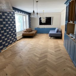 INSIDE HOME - DAMIAN RUSINEK - Salon w trakcie aranżacji: parkiet ułożony w jodełkę, niebieska zabudowa meblowa, tapeta w ważki, dwie sofy w różnych kolorach, designerskie lampy wiszące.