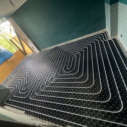 INSIDE HOME - DAMIAN RUSINEK - Układanie ogrzewania podłogowego: białe rury ułożone w pętle na czarnej macie izolacyjnej, widok z góry w trakcie montażu, surowy stan wykończenia ścian i sufitu.