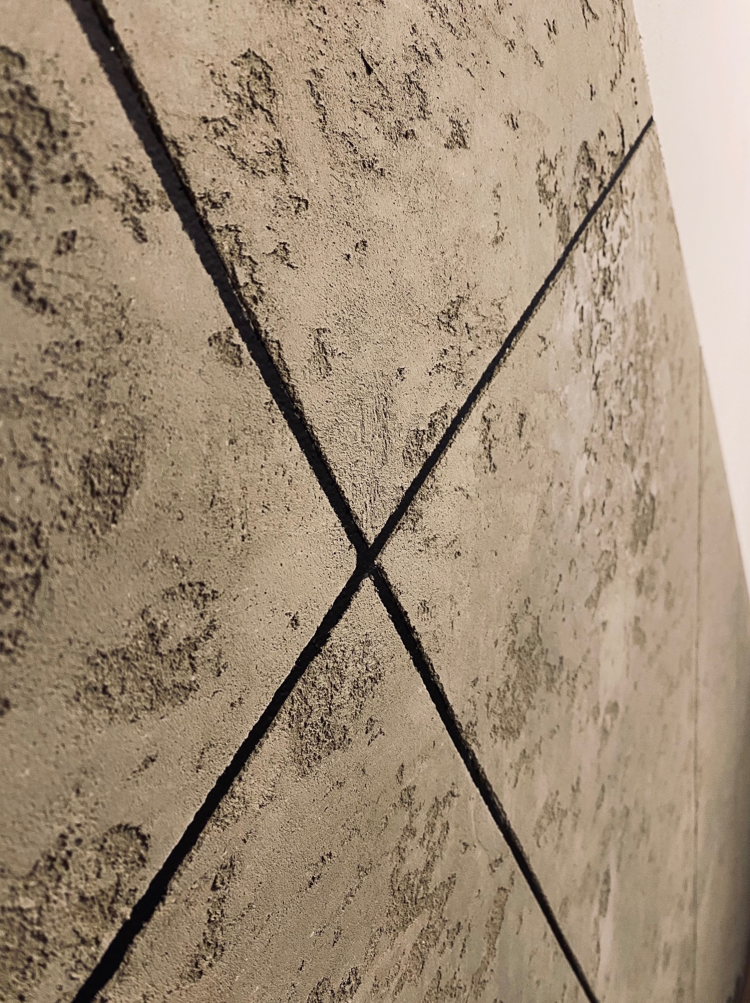 Ściana wykończona płytami z szarego betonu architektonicznego z widocznymi fugami krzyżującymi się na środku, faktura betonu z nierównościami i plamami, oświetlenie boczne.