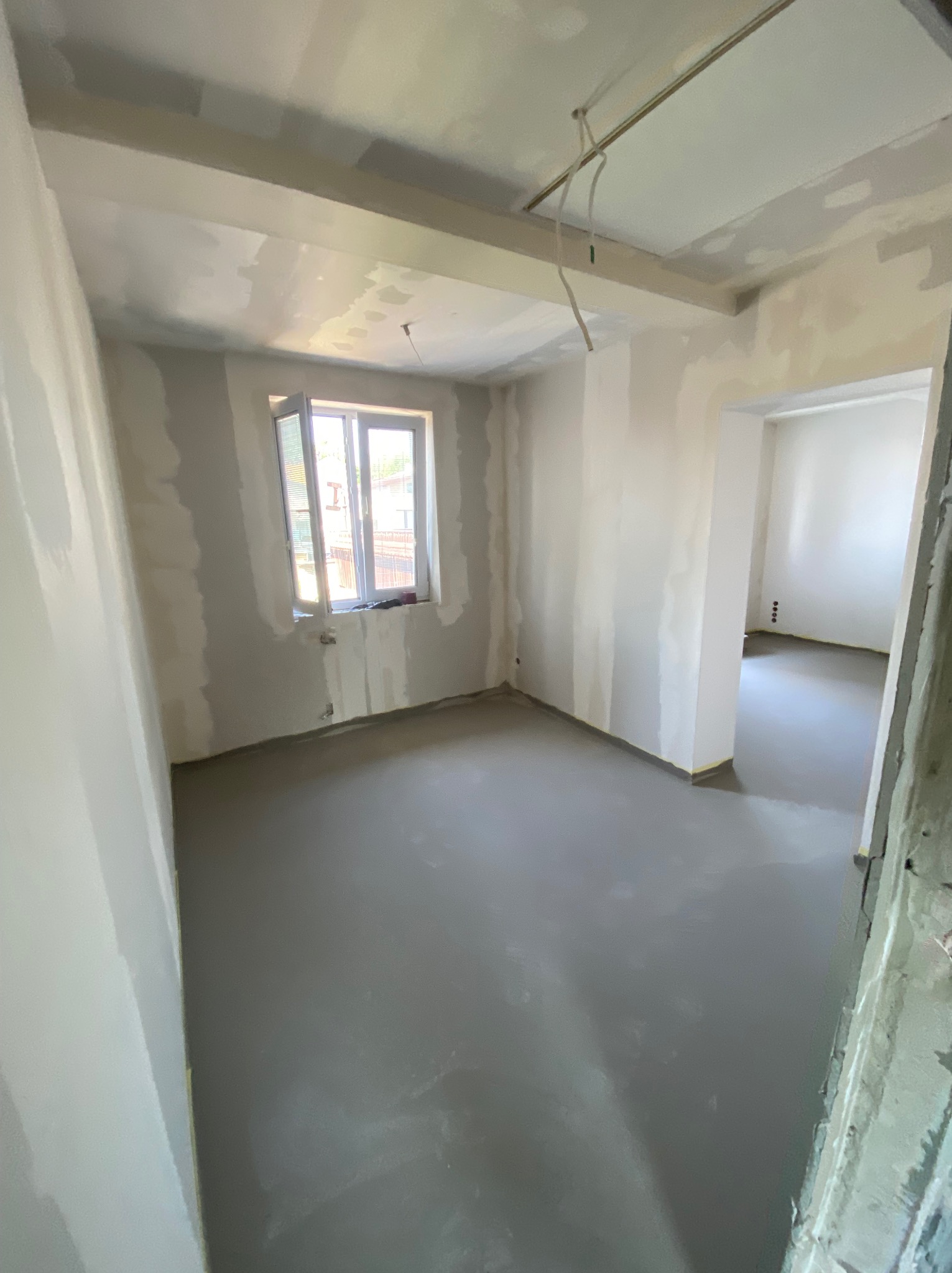 Surowe wnętrze w trakcie prac wykończeniowych: szpachlowane ściany, wylewka podłogowa, okno z widokiem na zewnątrz, widoczne kable pod sufitem.