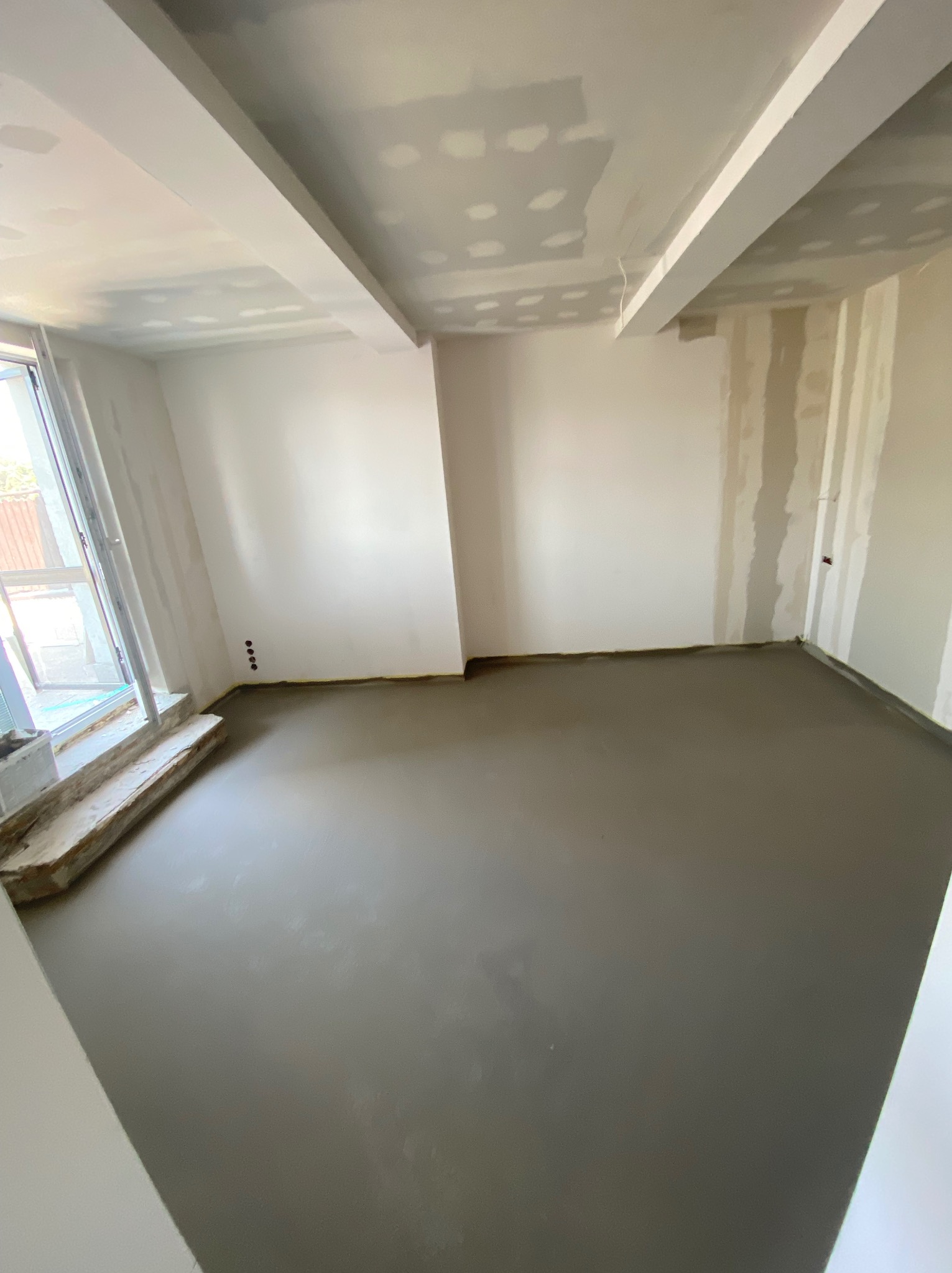 Surowe wnętrze w trakcie prac wykończeniowych: wylany beton na podłodze, ściany z fragmentami płyt kartonowo-gipsowych, sufit z widocznymi łączeniami i profilem, okno z surowym parapetem.