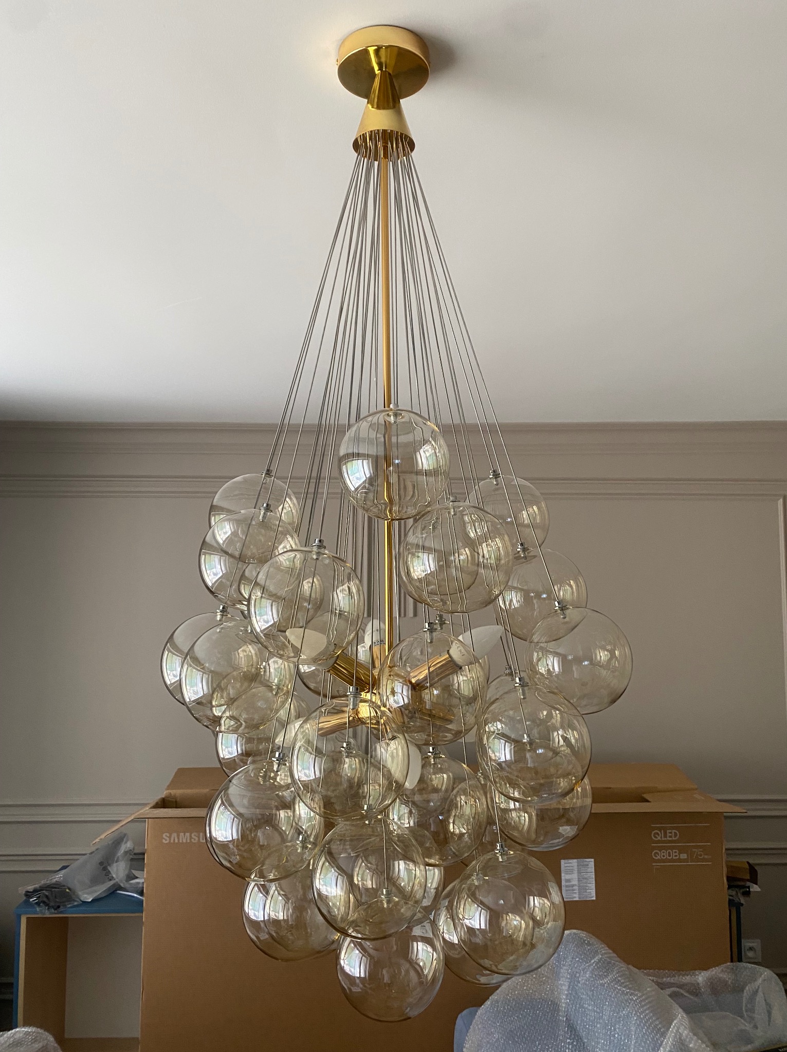 Złoty żyrandol z kulistymi, przezroczystymi kloszami zawieszony na cienkich linkach, widok od dołu, w tle kartonowe pudło z telewizorem Samsung QLED