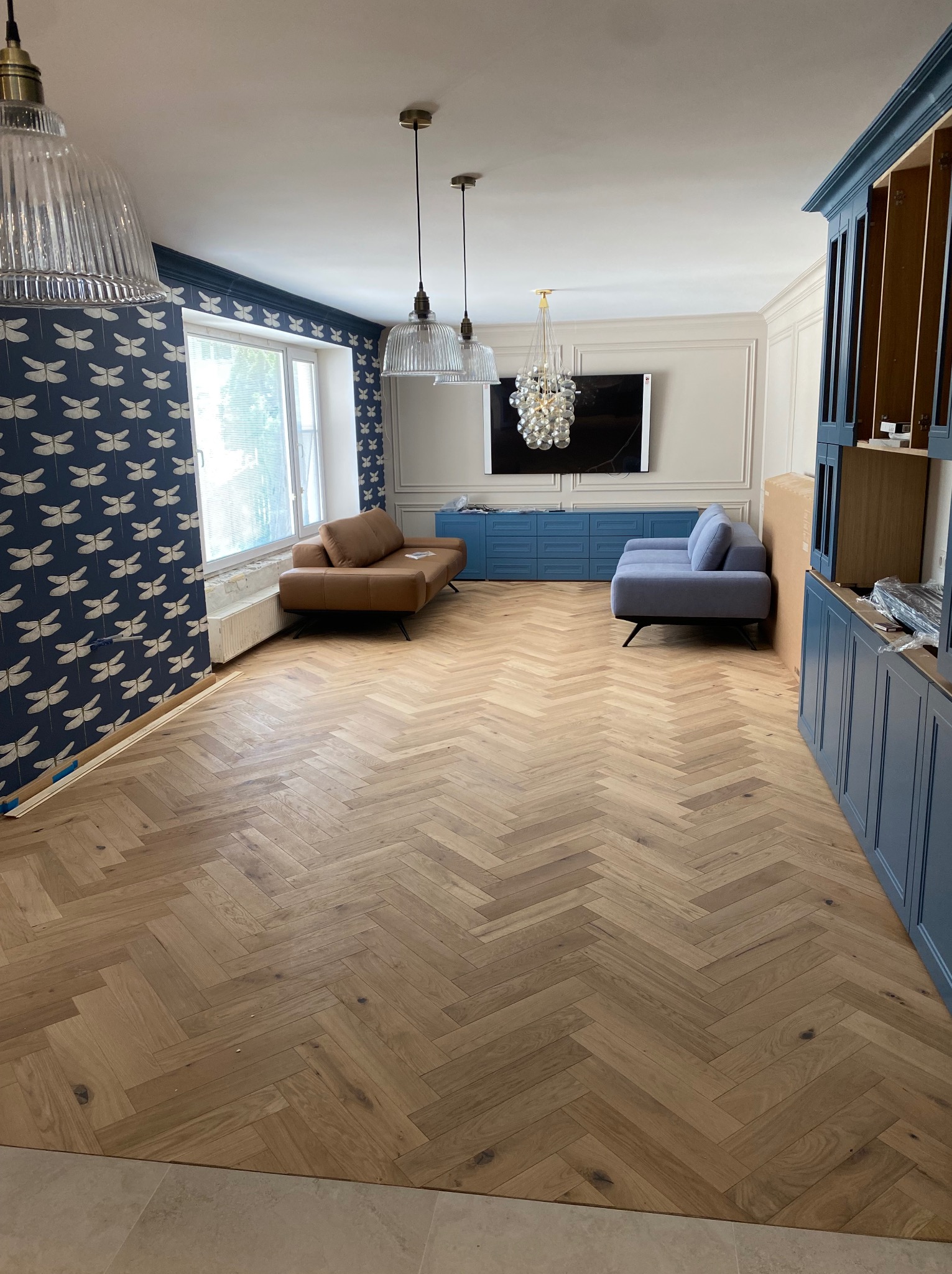 Salon w trakcie aranżacji: parkiet ułożony w jodełkę, niebieska zabudowa meblowa, tapeta w ważki, dwie sofy w różnych kolorach, designerskie lampy wiszące.