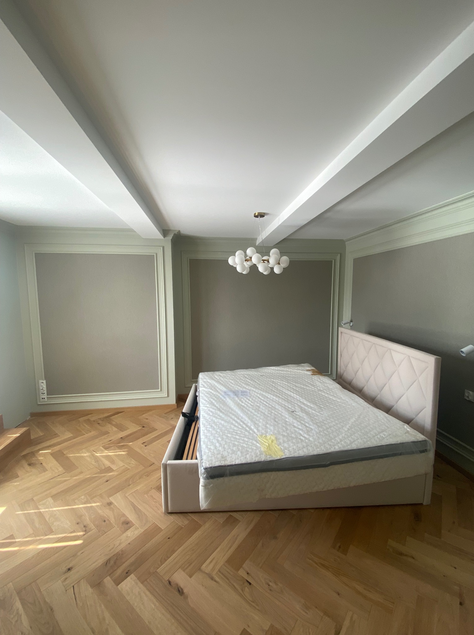 Jasny pokój z parkietem w jodełkę, łóżko z tapicerowanym zagłówkiem w folii, ozdobne listwy ścienne i nowoczesna lampa sufitowa.