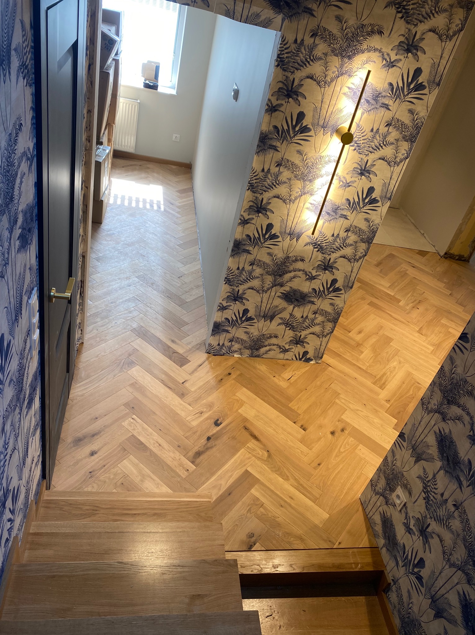 Ukończony remont korytarza z drewnianą podłogą ułożoną w jodełkę, schodami i ścianami z tapetą w palmy oraz nowoczesnym oświetleniem.