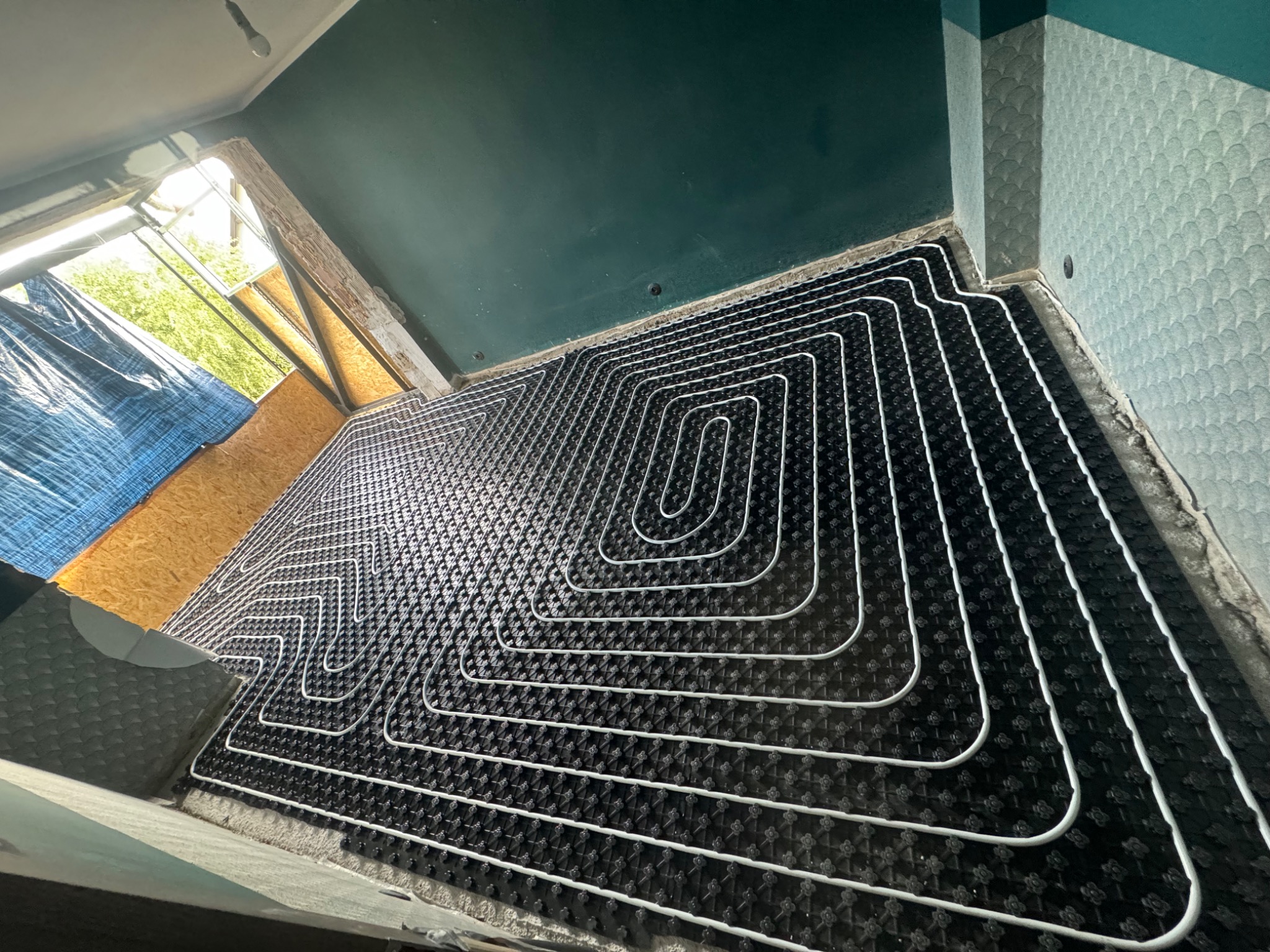 Układanie ogrzewania podłogowego: białe rury ułożone w pętle na czarnej macie izolacyjnej, widok z góry w trakcie montażu, surowy stan wykończenia ścian i sufitu.