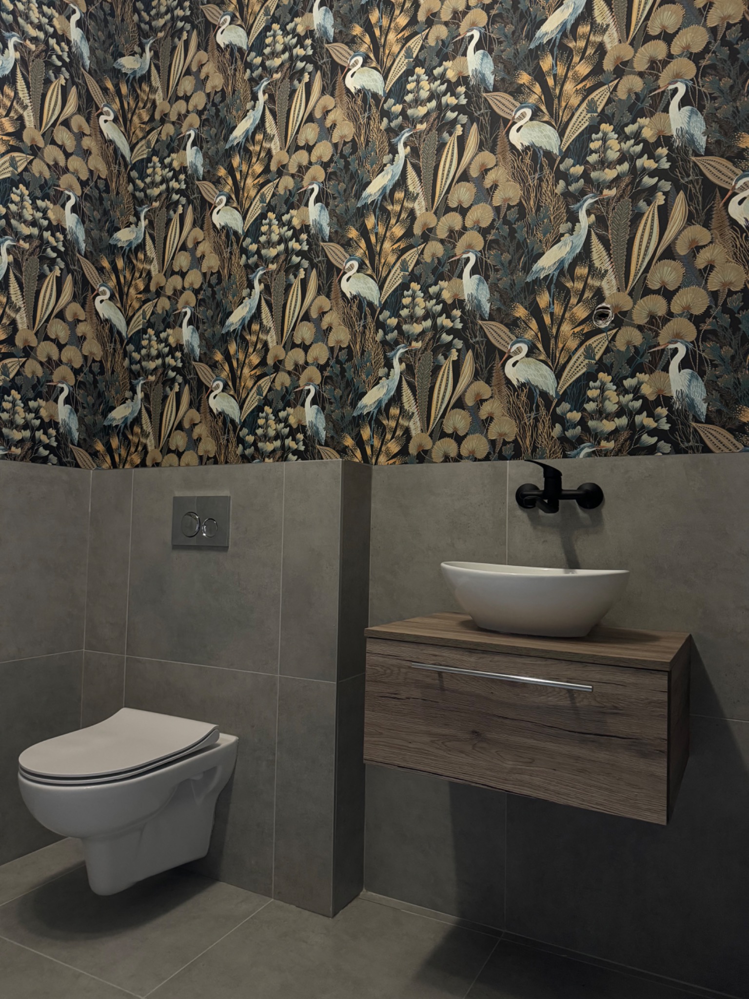 Nowoczesna łazienka z wiszącą toaletą, umywalką nablatową na szafce z drewnopodobnym dekorem i tapetą w motywy roślinne i ptasie.