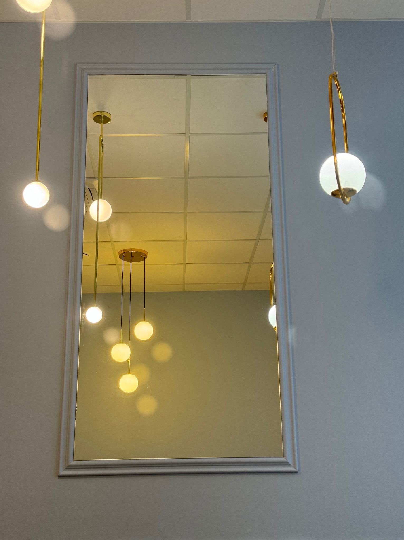 Lustro w szarej ramie na szarej ścianie, odbijające nowoczesne lampy kuliste z elementami w kolorze złotym, wiszące na białym suficie.