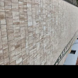 Długa ścieżka wyłożona beżowo-brązową kostką o nieregularnym wzorze, widoczna perspektywa z lekkiego ukosa, z zielonym wężem ogrodowym wzdłuż krawędzi i fragmentem budynku w tle.