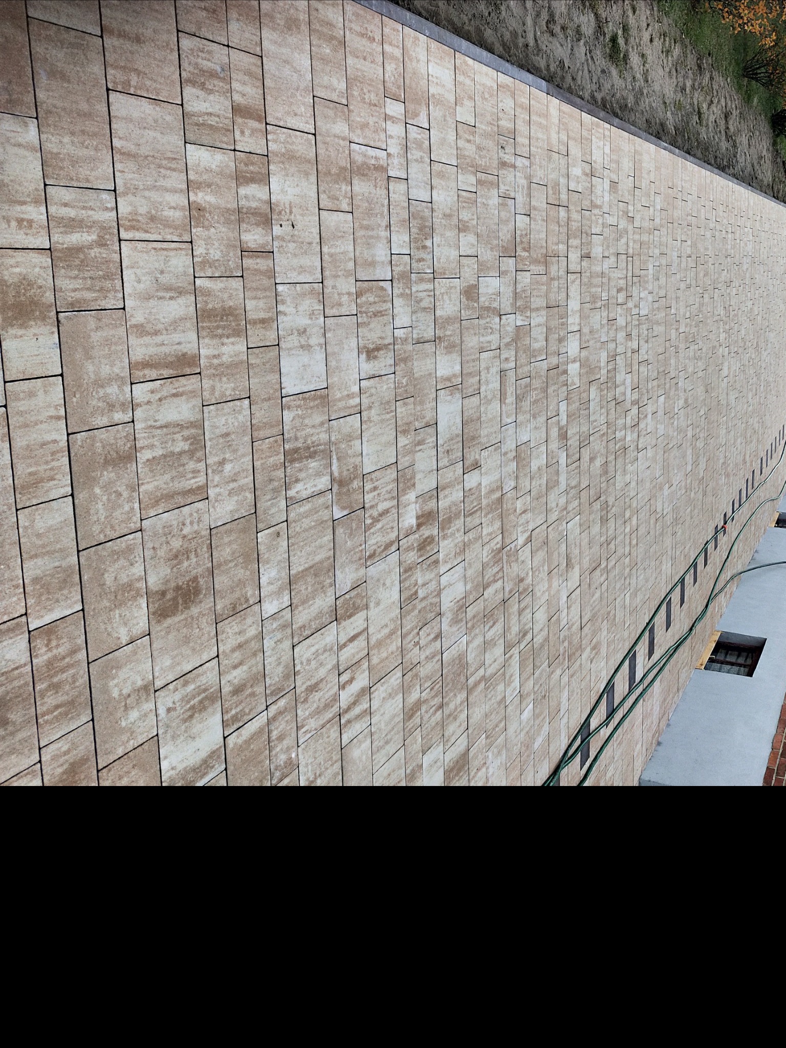 Długa ścieżka wyłożona beżowo-brązową kostką o nieregularnym wzorze, widoczna perspektywa z lekkiego ukosa, z zielonym wężem ogrodowym wzdłuż krawędzi i fragmentem budynku w tle.