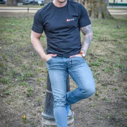 Uśmiechnięty mężczyzna w sportowej koszulce z tatuażami na ramieniu, oparty o latarnię w parku, prezentujący sylwetkę.