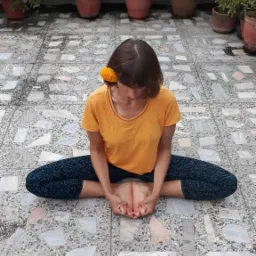 Kobieta w pomarańczowej koszulce siedzi na podłodze w pozycji motyla, z żółtym kwiatem we włosach, na tle tarasu z doniczkami.