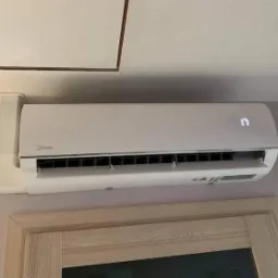 Biały klimatyzator Midea zamontowany na ścianie nad drewnianą ościeżnicą drzwi. Na wyświetlaczu widoczna temperatura 17 stopni.