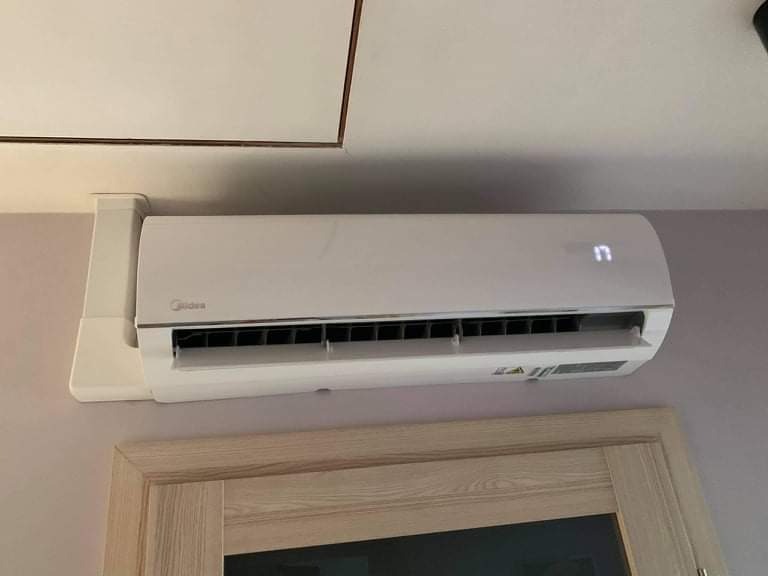 Biały klimatyzator Midea zamontowany na ścianie nad drewnianą ościeżnicą drzwi. Na wyświetlaczu widoczna temperatura 17 stopni.