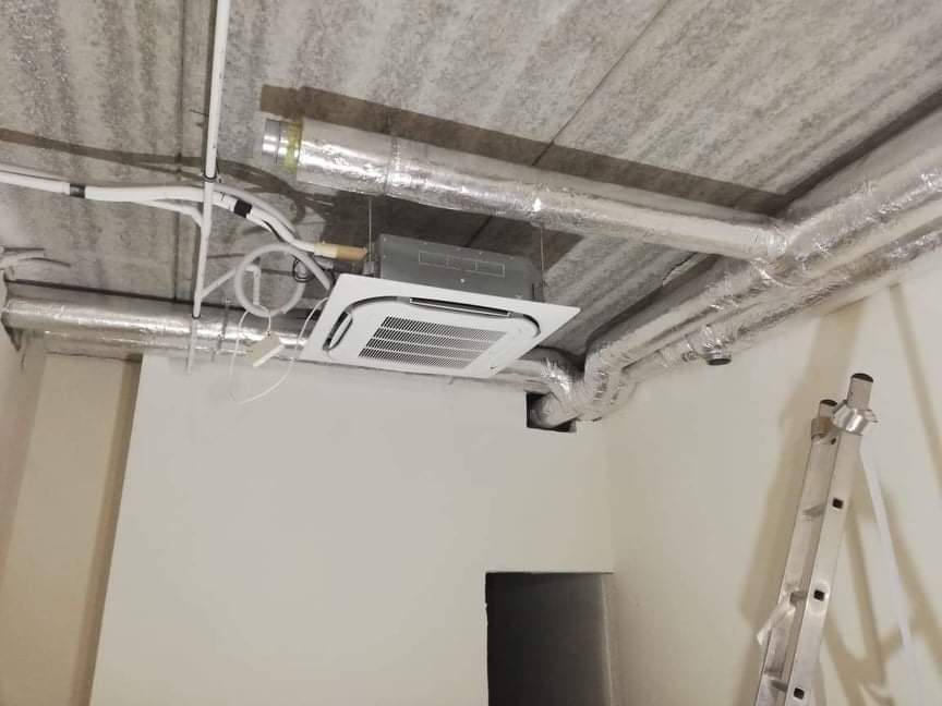 Kasetonowa jednostka klimatyzacyjna zamontowana w surowym wnętrzu biura, widoczne rury i instalacja, drabina oparta o ścianę.