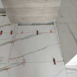 Układanie płytek podłogowych z wzorem marmuru, z wykorzystaniem systemu poziomowania, widoczny fragment drewnopodobnej podłogi.