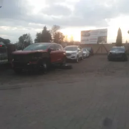 Uszkodzony czerwony samochód osobowy z widocznymi zniszczeniami karoserii stoi na placu, w tle zaparkowane inne pojazdy i billboard reklamowy.