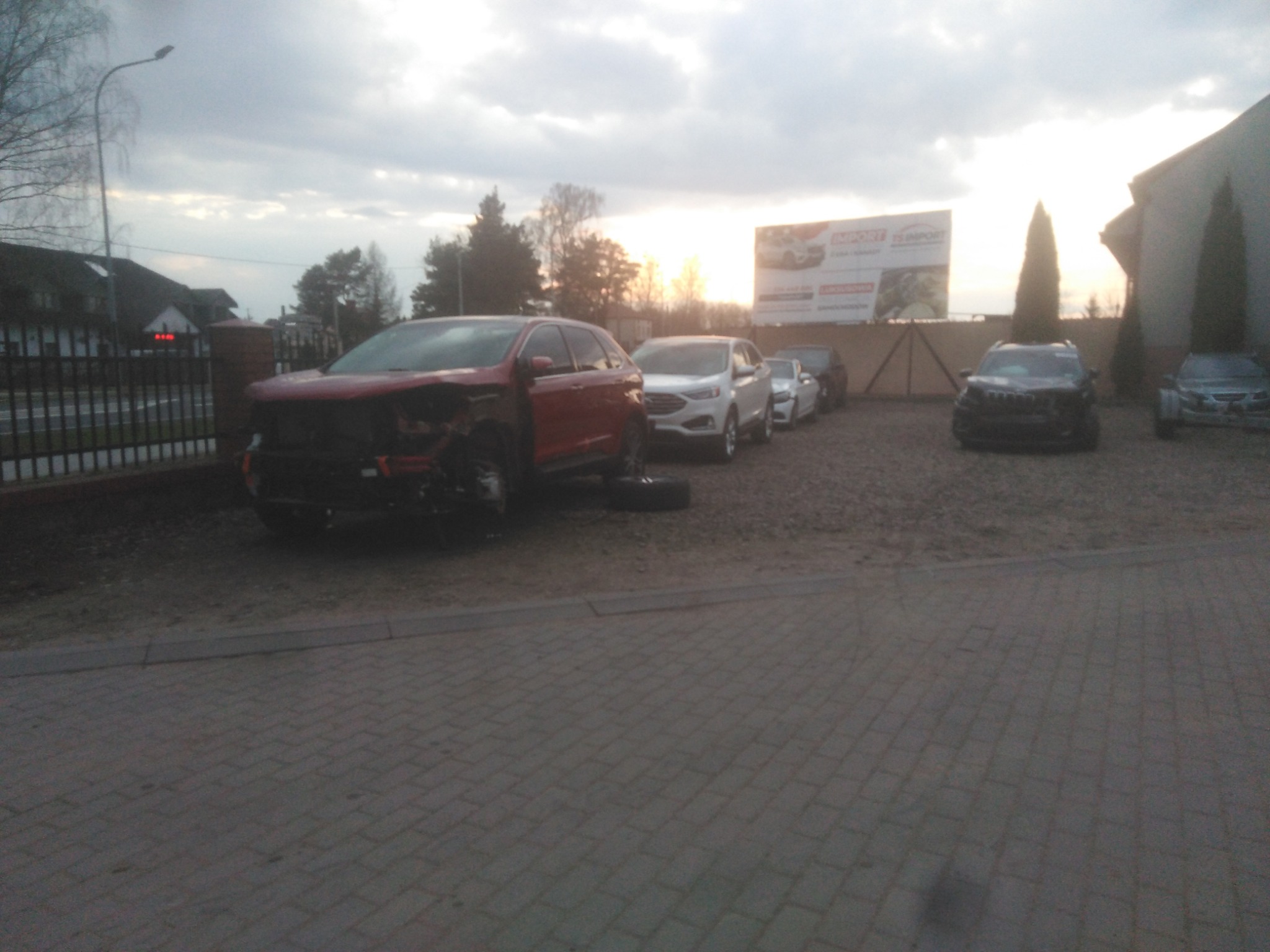 Uszkodzony czerwony samochód osobowy z widocznymi zniszczeniami karoserii stoi na placu, w tle zaparkowane inne pojazdy i billboard reklamowy.